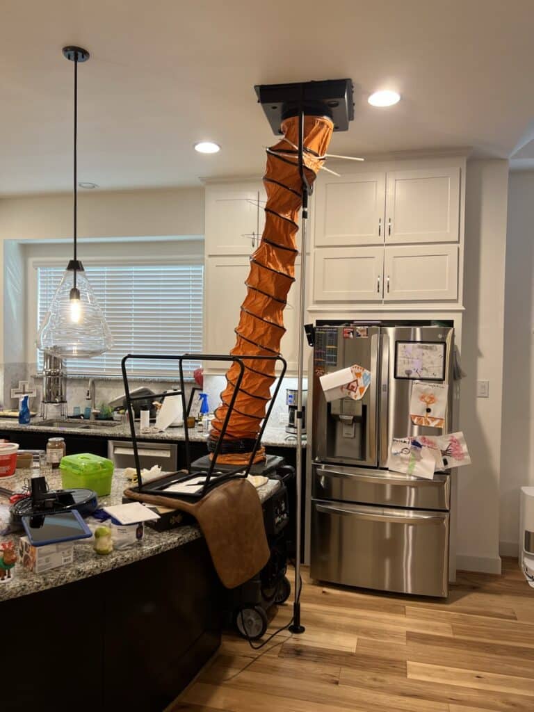 Professional air duct cleaning setup in a residential kitchen with NIKRO vacuum and orange hose connected to ceiling vent