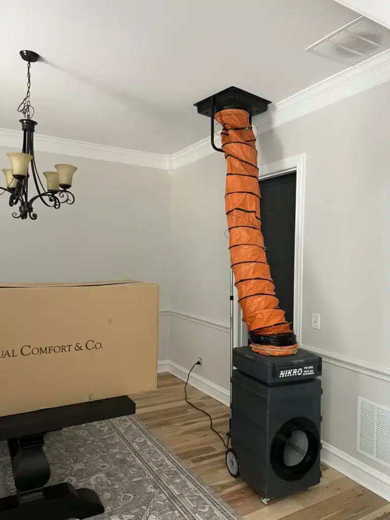 Air duct cleaning setup: HEPA air scrubber connected by orange ducting to a ceiling vent in a home