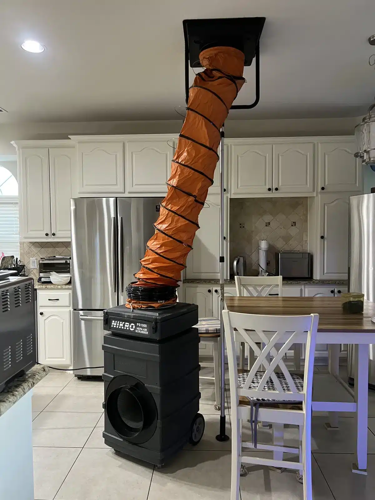 Air duct cleaning setup in a kitchen: ceiling vent connected by orange flex duct to a portable air scrubber