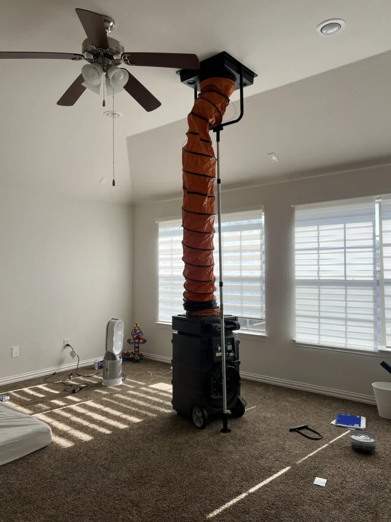 Air duct cleaning: flexible duct running from a floor machine to a ceiling vent in a carpeted room