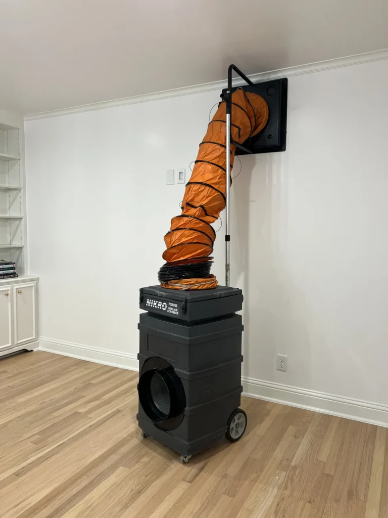 Air duct cleaning: HEPA air scrubber connected by orange flex duct to a return vent on a wood floor