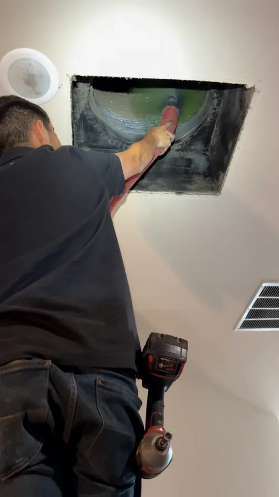 Air duct cleaning: HEPA air scrubber connected by orange duct to a ceiling opening in a dining room