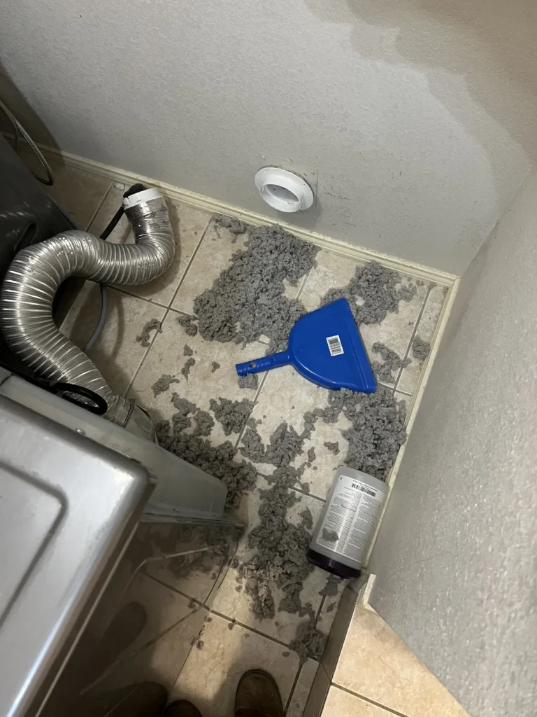 Dryer vent cleaning: lint clumps and a dustpan on the laundry room floor behind the dryer