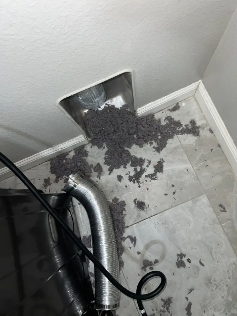 Dryer vent cleaning: lint spilling from the wall duct opening onto the floor behind the dryer