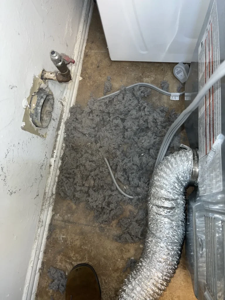 Dryer vent cleaning: lint buildup on the floor behind a pulled-out dryer, with flexible foil duct and gas line visible